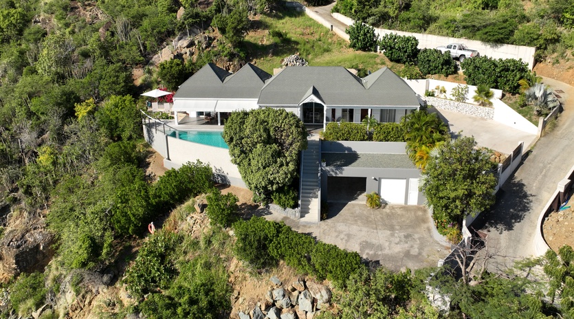 Aerial view of modern vacation villa surrounded by lush tropical vegetation, featuring swimming pool and private parking area.