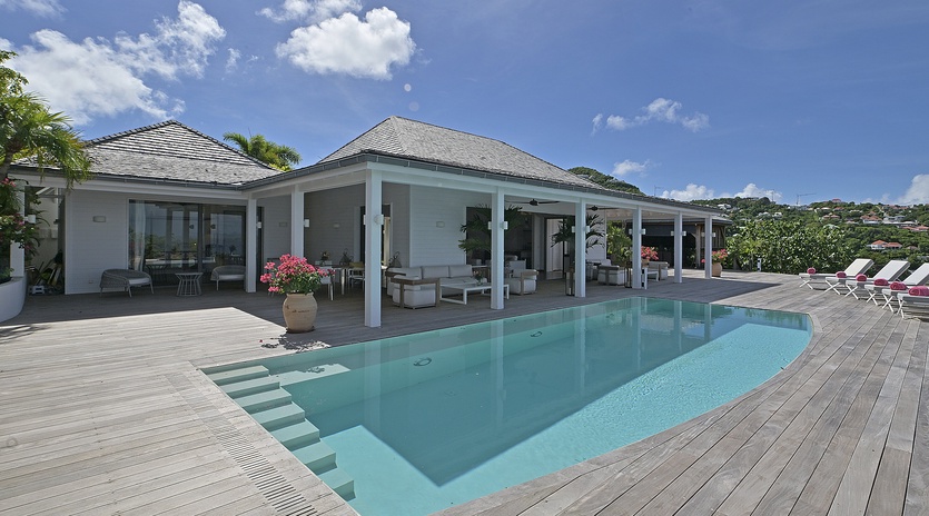 A contemporary villa showcases its sleek covered terrace and pristine swimming pool, surrounded by tropical landscaping under bright skies.