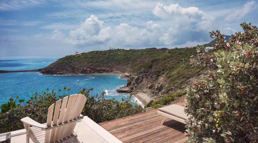 A secluded coastal hillside offers a dramatic panoramic view of turquoise waters, rugged cliffs, and lush green vegetation stretching toward distant villas under a Caribbean sky.