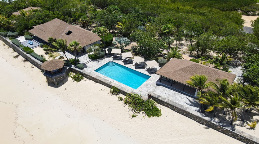 Aerial view showcasing a beachfront vacation property with private pool, multiple structures, and lush tropical landscaping set along pristine white sand and dense foliage.