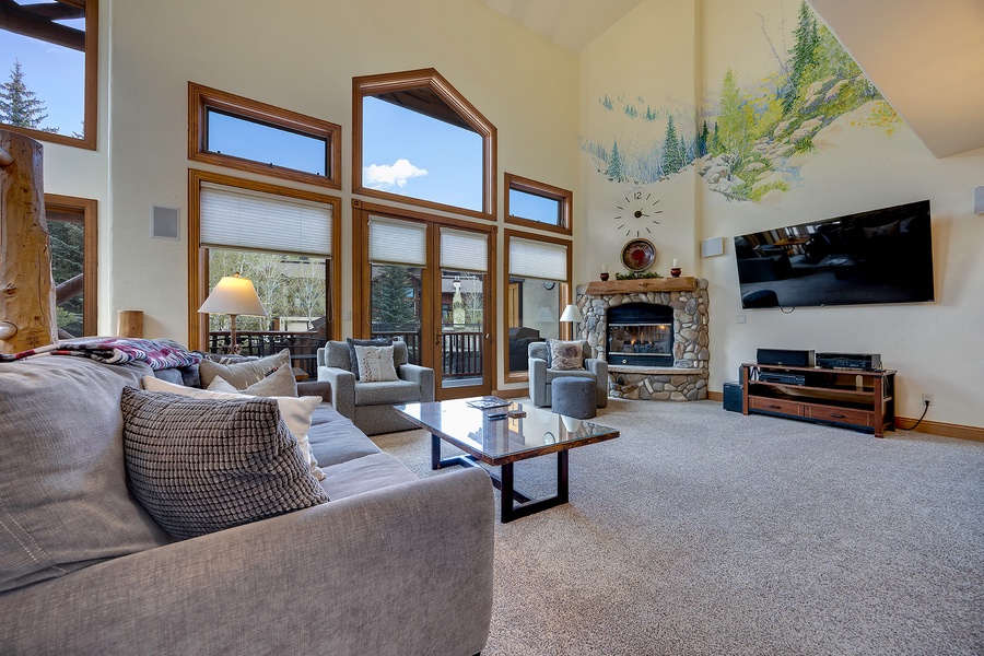 Sink into plush seating where soaring windows frame mountain views, and gather by the stone fireplace as sunlight streams through vaulted ceilings—your alpine retreat awaits.