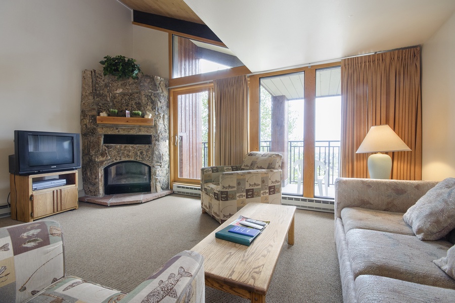 Relax in your bright, vaulted living room with a cozy stone fireplace, plush seating, and balcony access framing peaceful mountain and forest views.