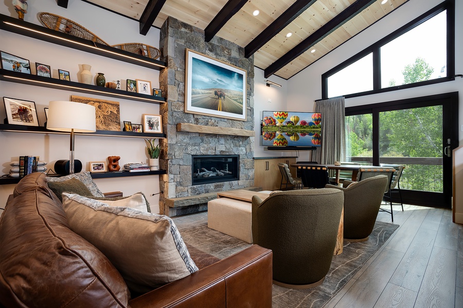 Unwind in your inviting living room featuring a stone fireplace, leather seating, and stunning forest views through floor-to-ceiling windows—your perfect mountain retreat.