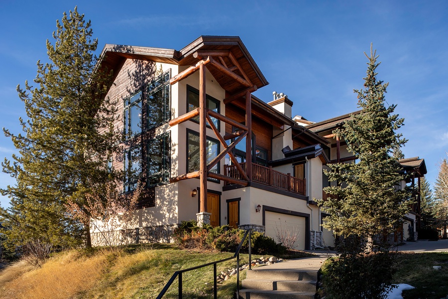 This alpine-inspired mountain retreat features a striking multi-level design with wood and stone elements, nestled among tall evergreens under clear blue skies.