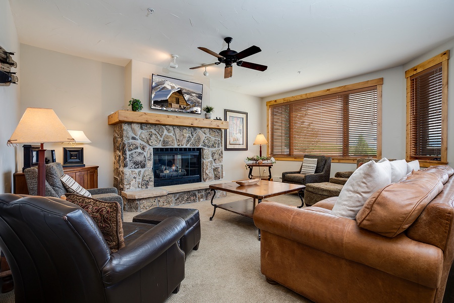 Unwind in your inviting living room with cozy leather seating, a warm stone fireplace, and mountain views through natural wood blinds creating the perfect alpine retreat.