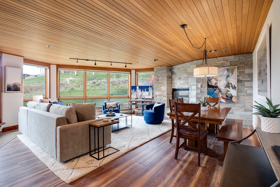 Unwind in this sun-drenched living space where rich wood tones meet modern comfort. Sink into plush seating, enjoy hillside views through expansive windows, and gather at the dining table beneath the warmth of natural wood ceilings.