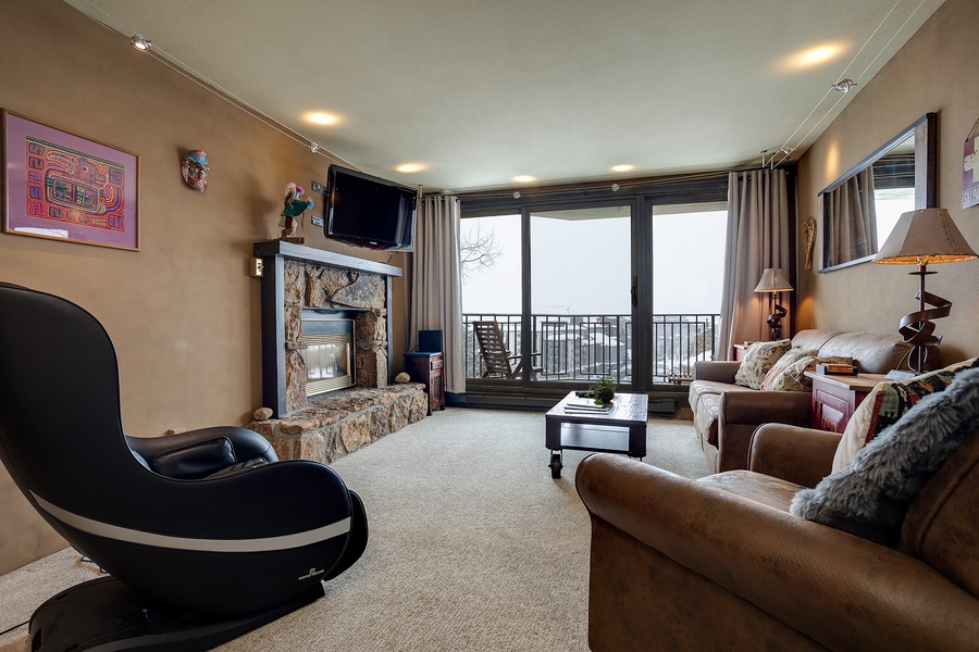 Your inviting living space features plush sofas, a cozy stone fireplace, and a private balcony where you can unwind while enjoying scenic mountain views through floor-to-ceiling windows.