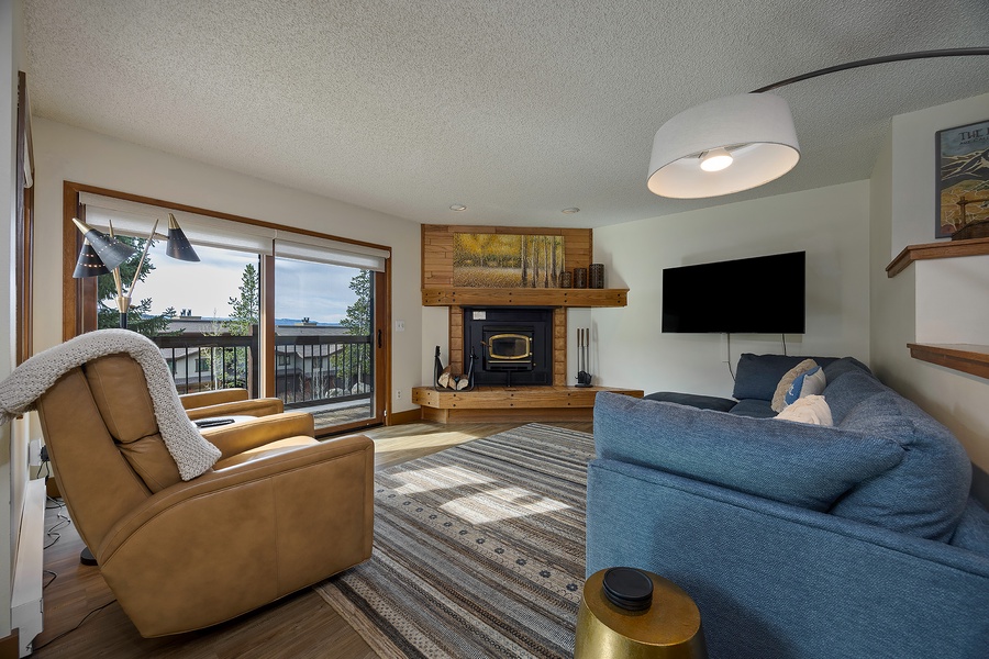 Settle into your cozy living room where sunlight streams through sliding glass doors, revealing forest and lake views. Unwind by the wood-burning fireplace or watch shows on the flat-screen TV.