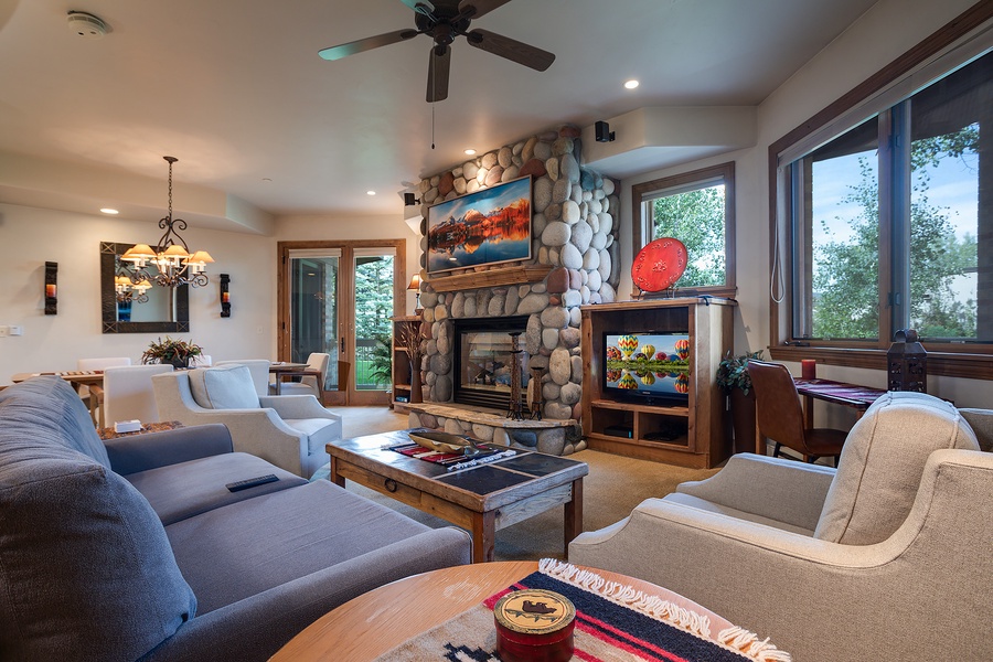 Relax in your spacious living room featuring a stunning stone fireplace, ceiling fan, and picture windows framing beautiful treetop views. Thoughtfully furnished with plush seating and warm lighting for effortless comfort.