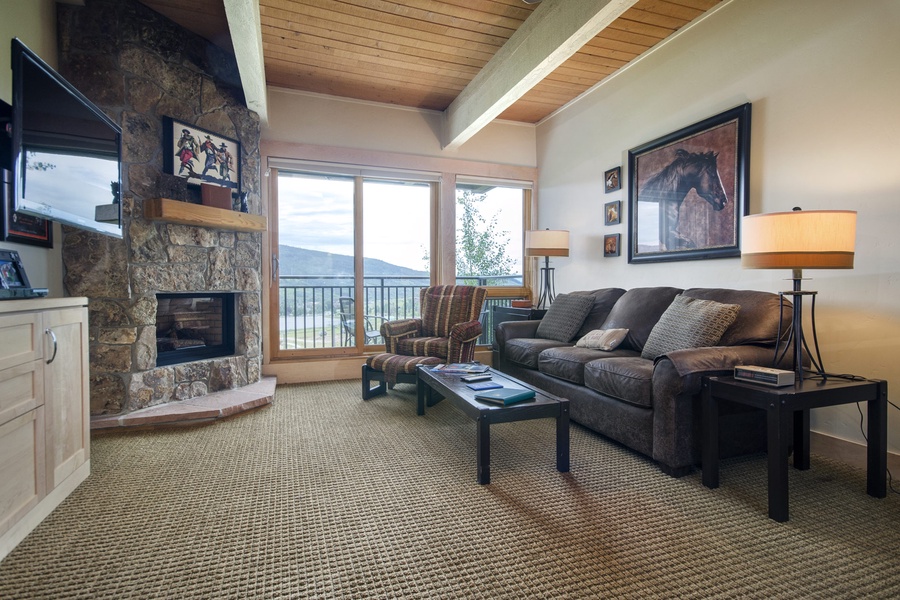 Sink into the plush sofa by the stone fireplace and enjoy stunning mountain views through expansive windows. The warm wood ceiling and cozy seating create your perfect retreat after a day exploring.