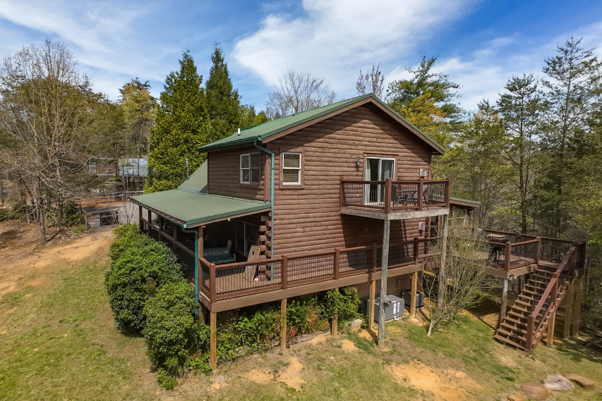 Elevated log cabin nestled among mature trees with wraparound deck and multi-level design in a peaceful wooded setting.