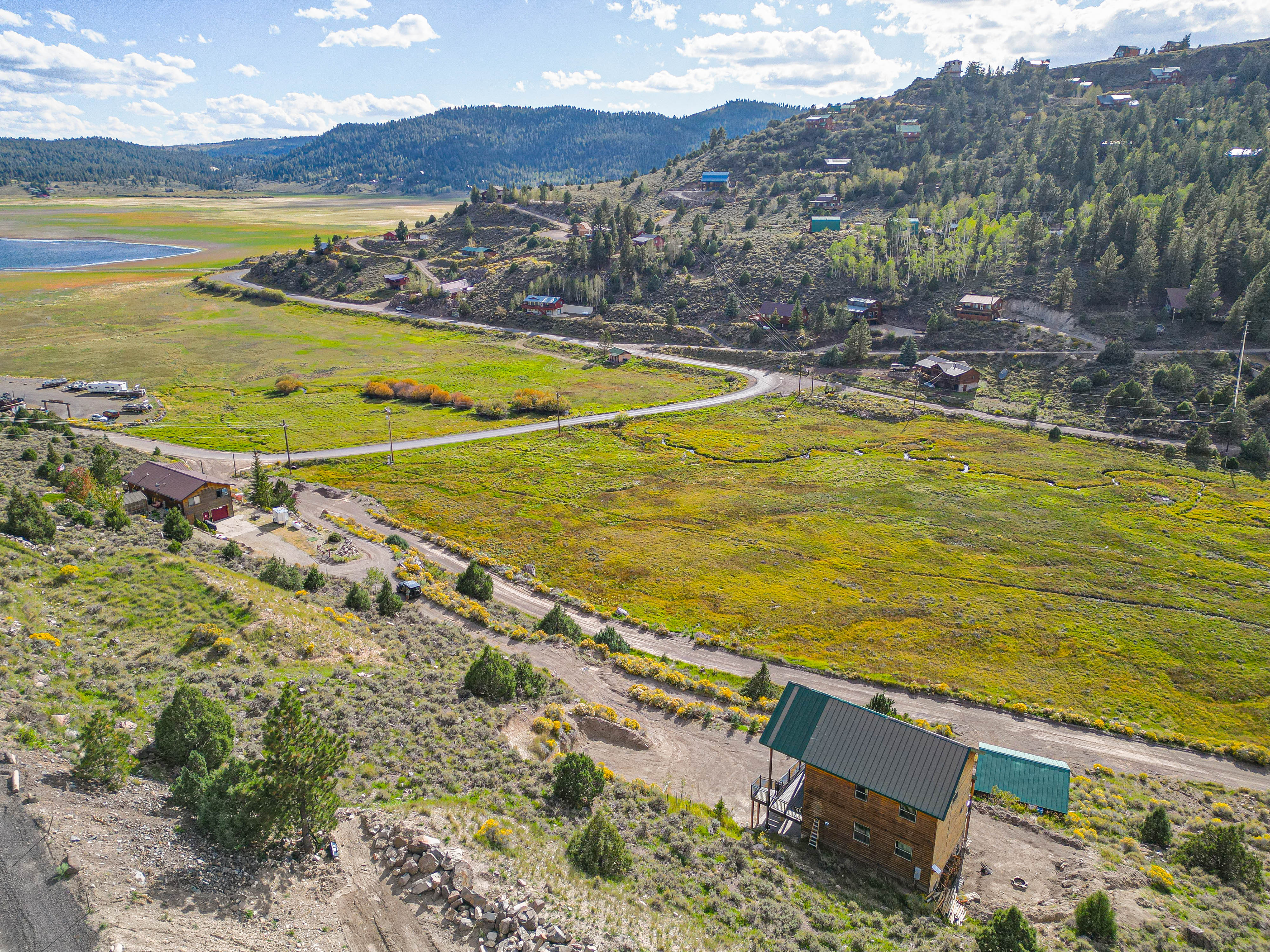 Aerial perspective of the property nestled in a scenic mountain valley with forested hillsides, open meadows, and a sparkling lake nearby.