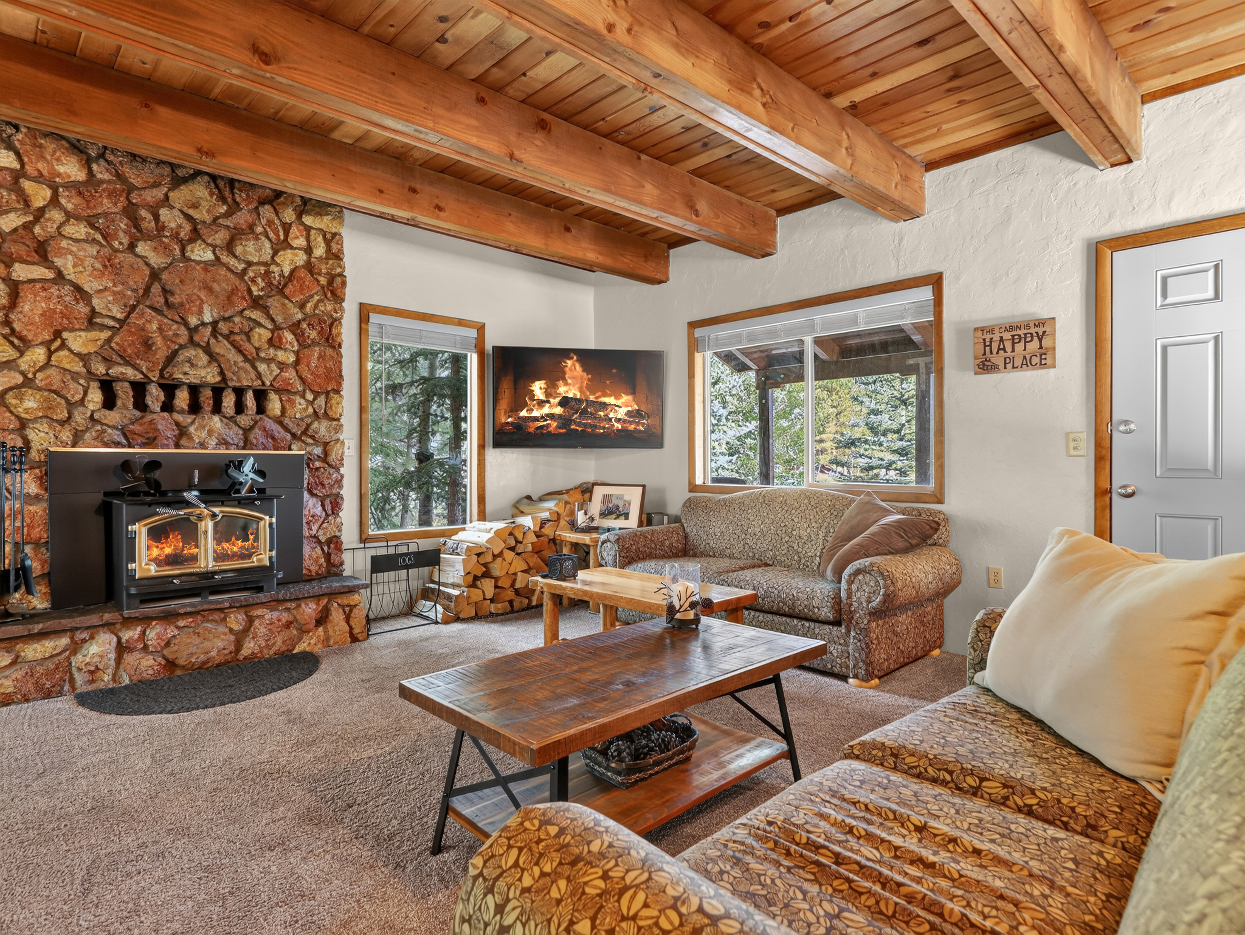 Warm up in your cozy living room featuring rustic wood-beam ceilings, a stunning stone fireplace, and peaceful wooded views through large windows. The inviting space includes comfortable seating and natural touches throughout.