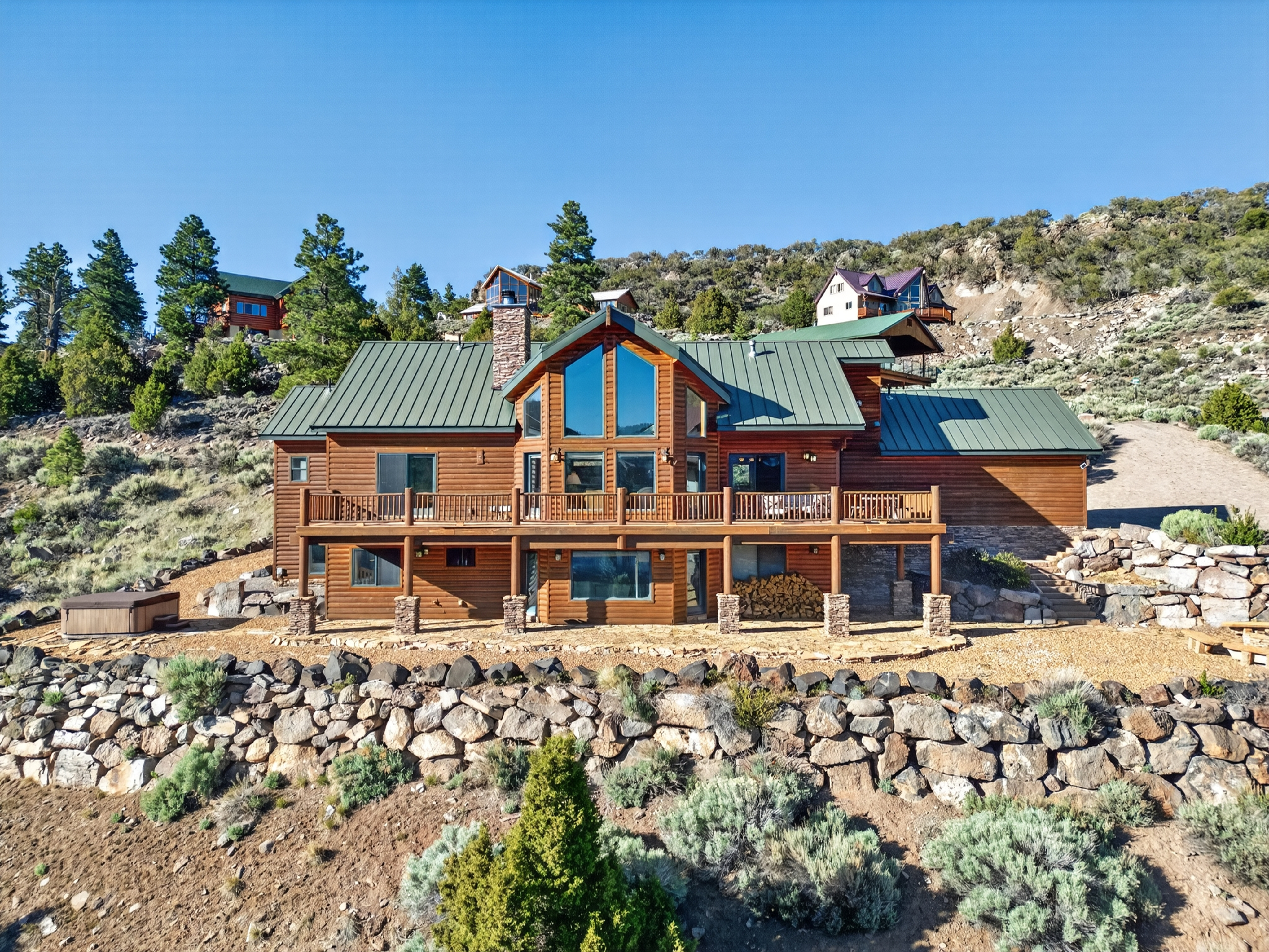 A striking mountain lodge nestled on a hillside, surrounded by evergreens and rugged terrain. The multi-level log home features expansive windows, wraparound decking, and stone accents, offering a peaceful retreat in a scenic natural setting.