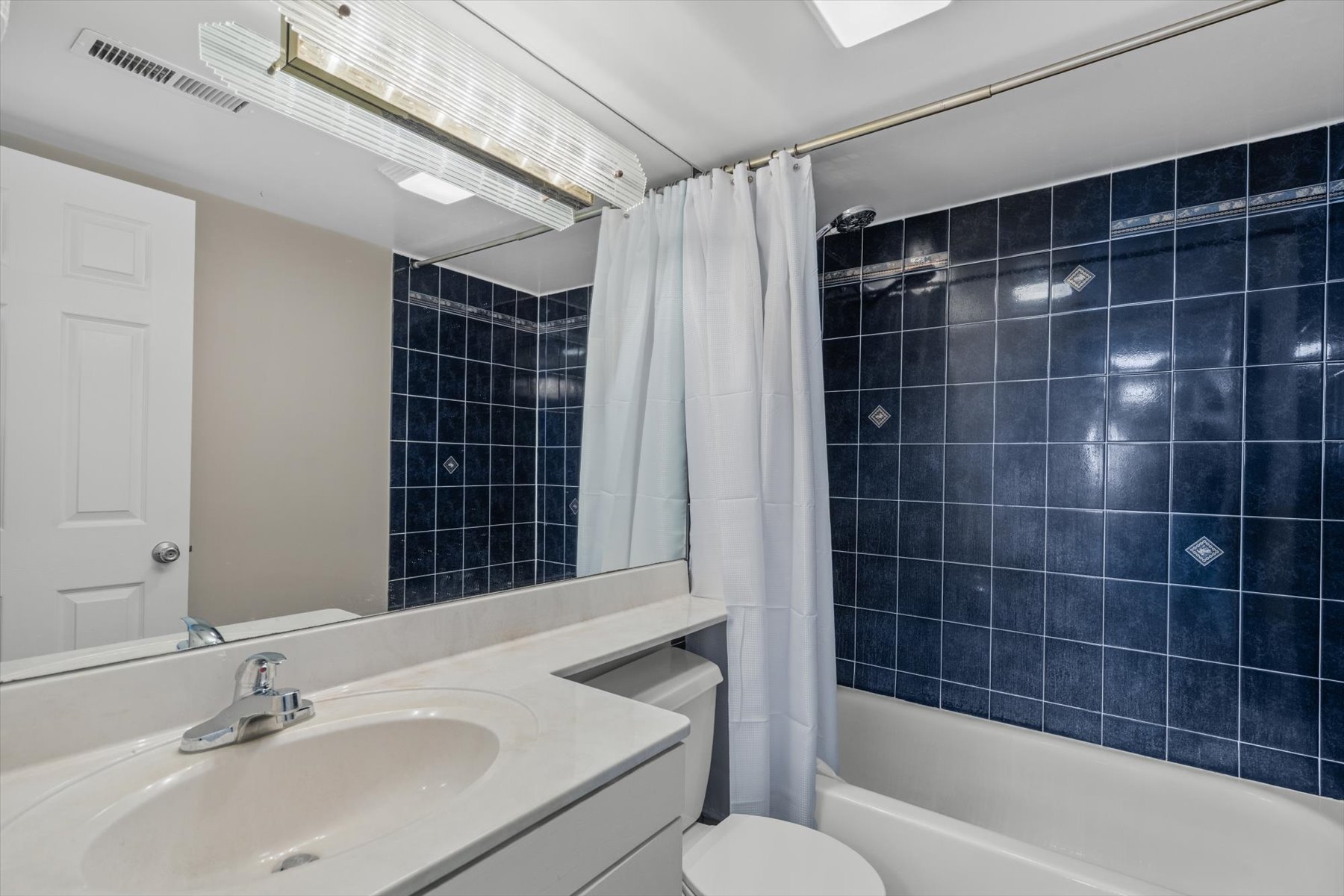 This clean bathroom features a spacious white vanity, a large mirror, and bright lighting right next to the bath area.