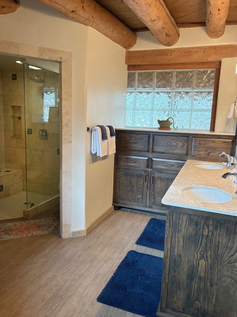 Step into your private bathroom sanctuary with stunning wood beam ceilings, a spacious glass-enclosed shower, and rustic charm. Soft towels and natural light through decorative glass blocks create a warm, relaxing retreat.