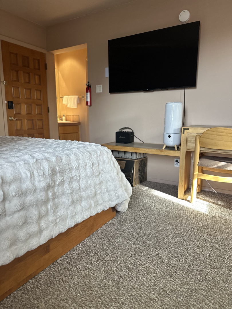 Unwind in your cozy bedroom with plush bedding, large wall-mounted TV, and thoughtful workspace setup for ultimate comfort and convenience.