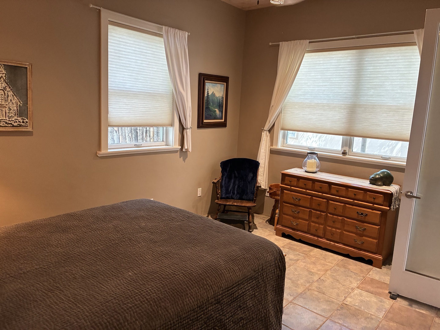 Your peaceful bedroom retreat features a plush bed and warm natural light. Unwind in the cozy rocking chair or unpack in the spacious wooden dresser, with soft curtains framing views of the wintery outdoors.