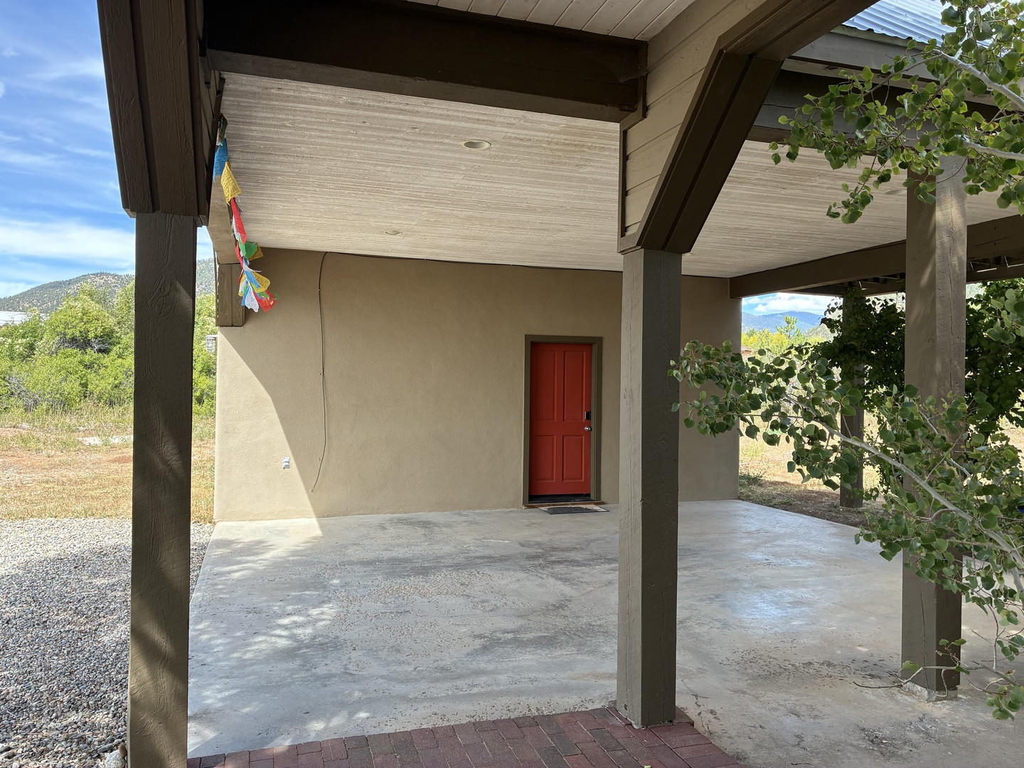 Your arrival is marked by a covered entrance with distinctive timber beams and a welcoming red door, set against a peaceful natural landscape and mountain backdrop.