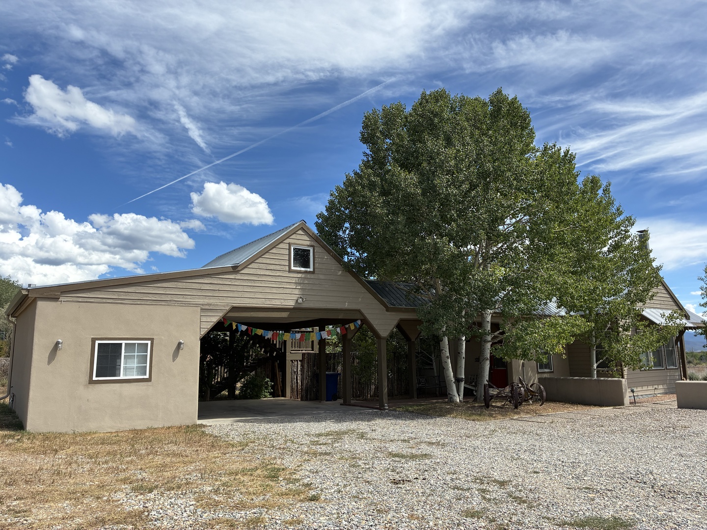 The property sits on spacious grounds with ample gravel parking under a covered carport, sheltered by tall, mature trees that offer natural shade and a sense of peaceful seclusion.