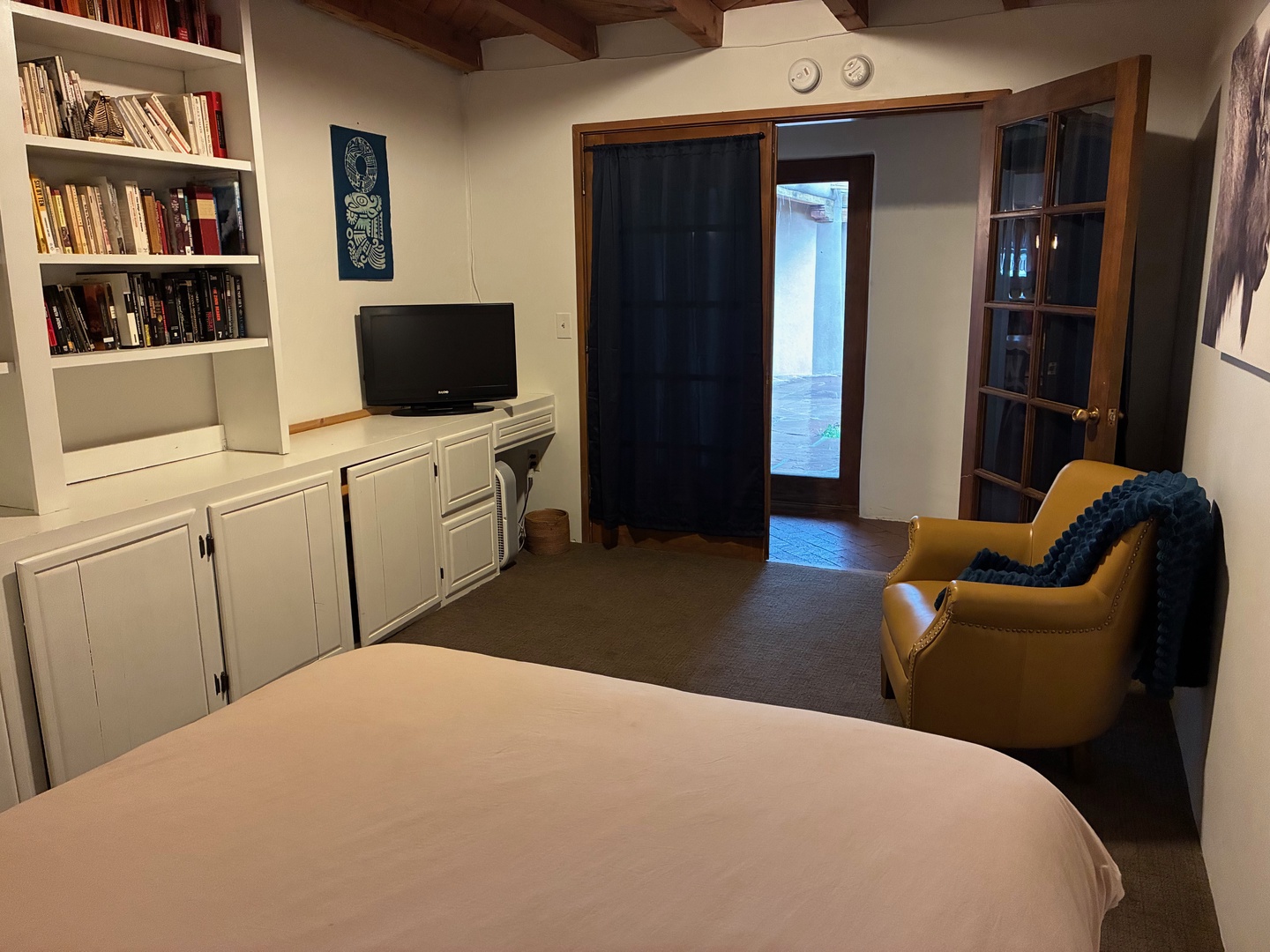 Your cozy retreat awaits with a plush bed, a warm reading nook by built-in bookshelves, and a comfortable armchair with a soft throw. The space opens to an outdoor courtyard, blending comfort with quiet charm.
