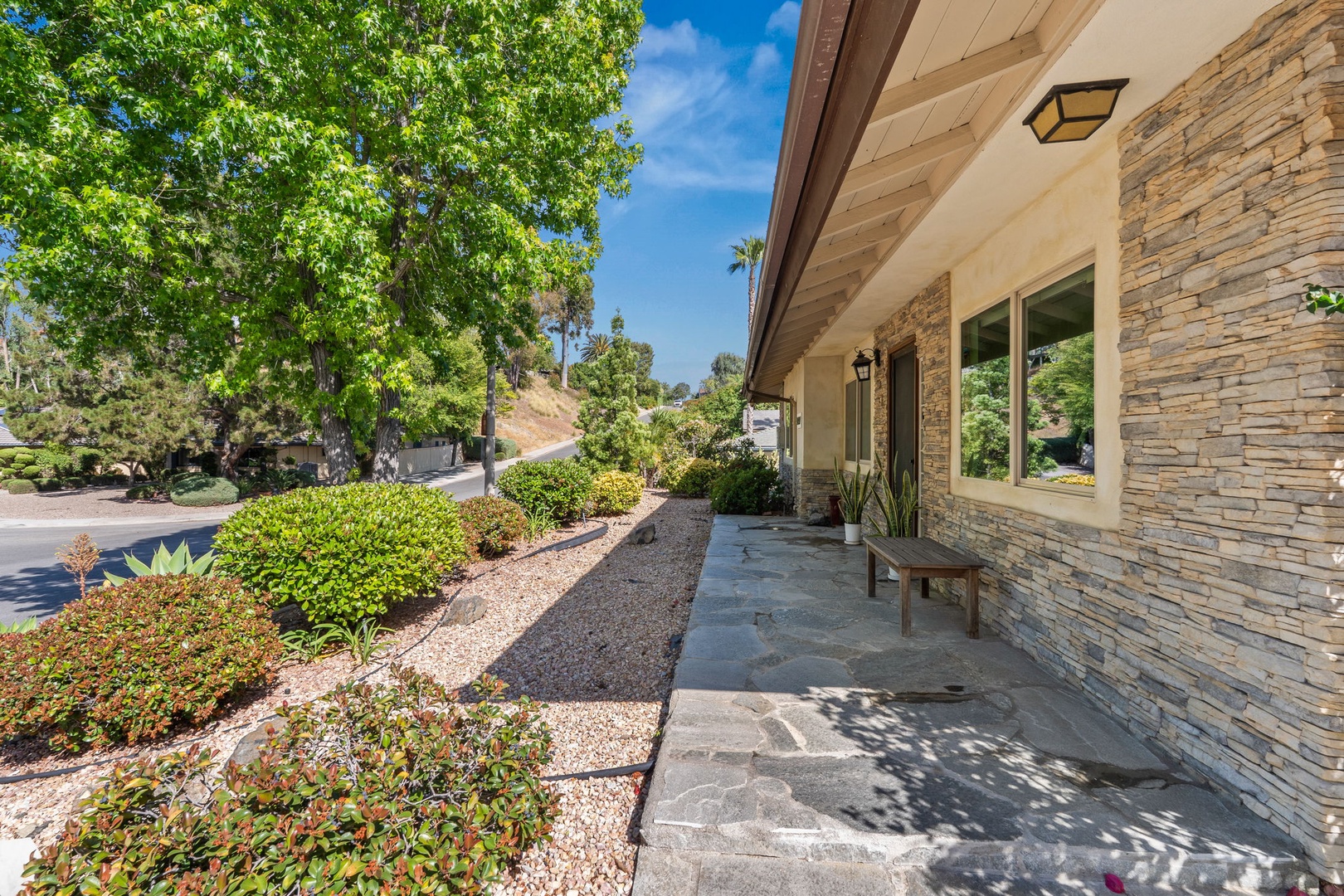 Modern stone facade entrance with landscaped gardens and shaded terrace seating area in a quiet residential neighborhood.