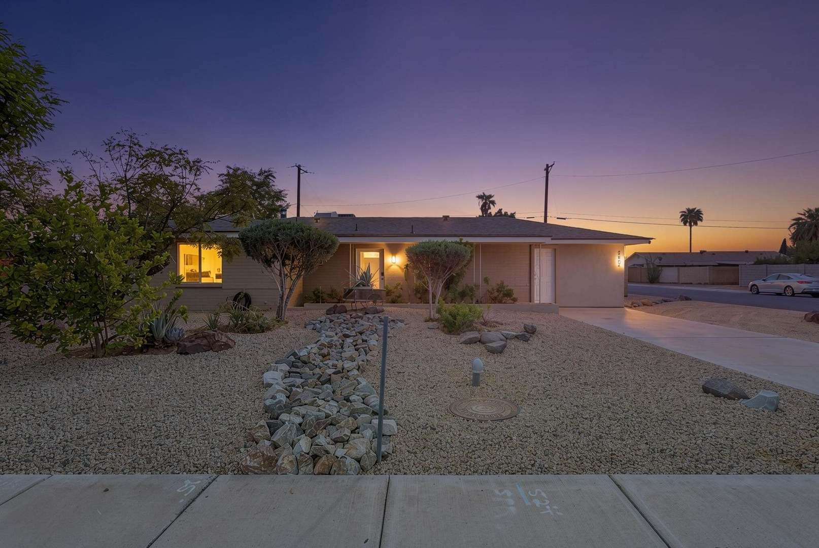 The property glows warmly against a stunning desert sunset, with desert landscaping and palm trees dotting the peaceful neighborhood.