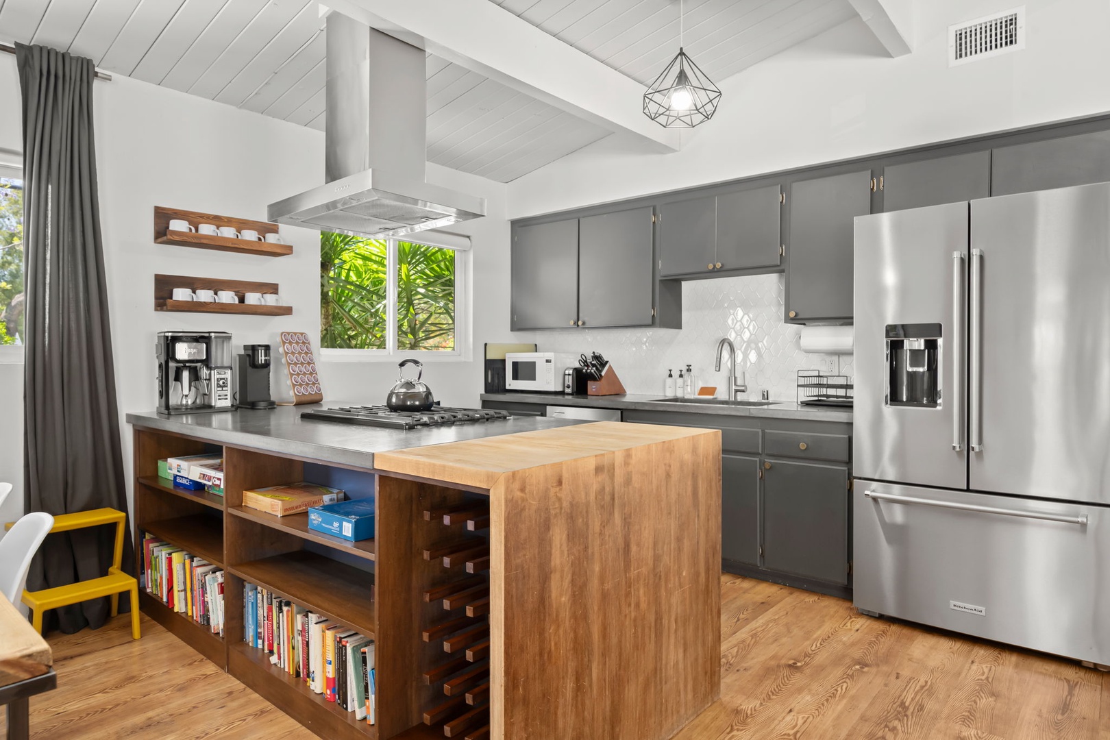 Your spacious kitchen features sleek gray cabinetry, stainless steel appliances, and a butcher-block island perfect for meal prep. Bright windows frame lush greenery, and open shelves invite your culinary creativity.