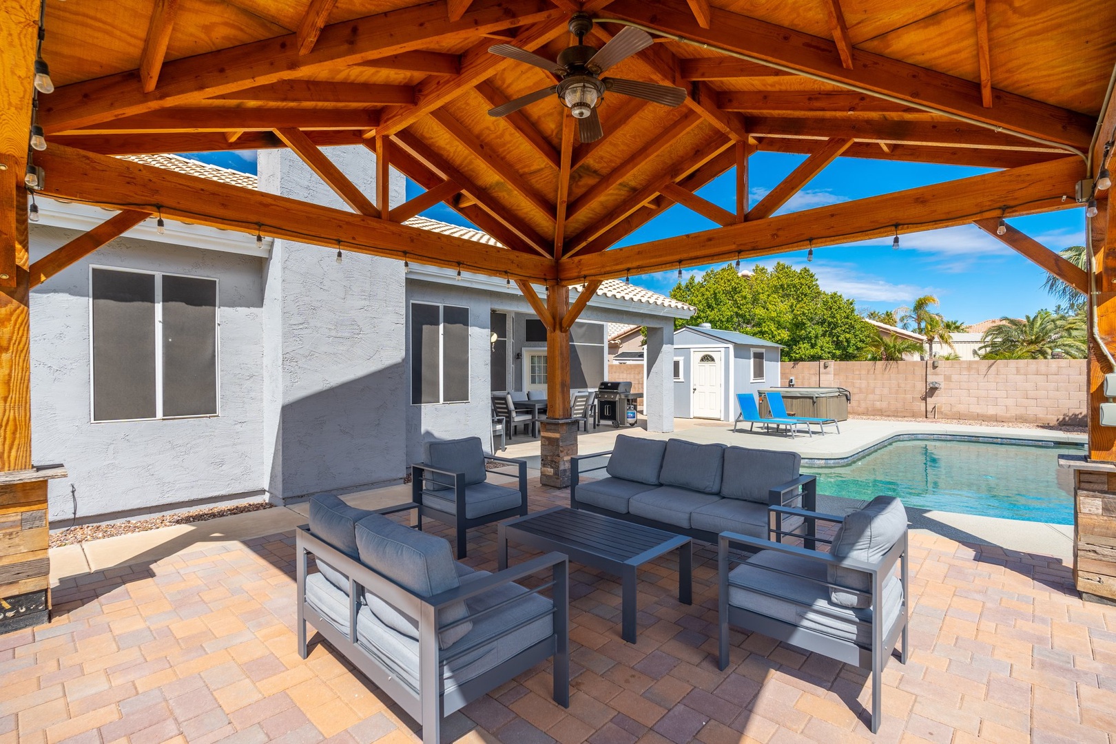 Relax under the vaulted wooden pergola with comfortable seating, a ceiling fan, and string lights, all overlooking your private pool and sunny backyard oasis.