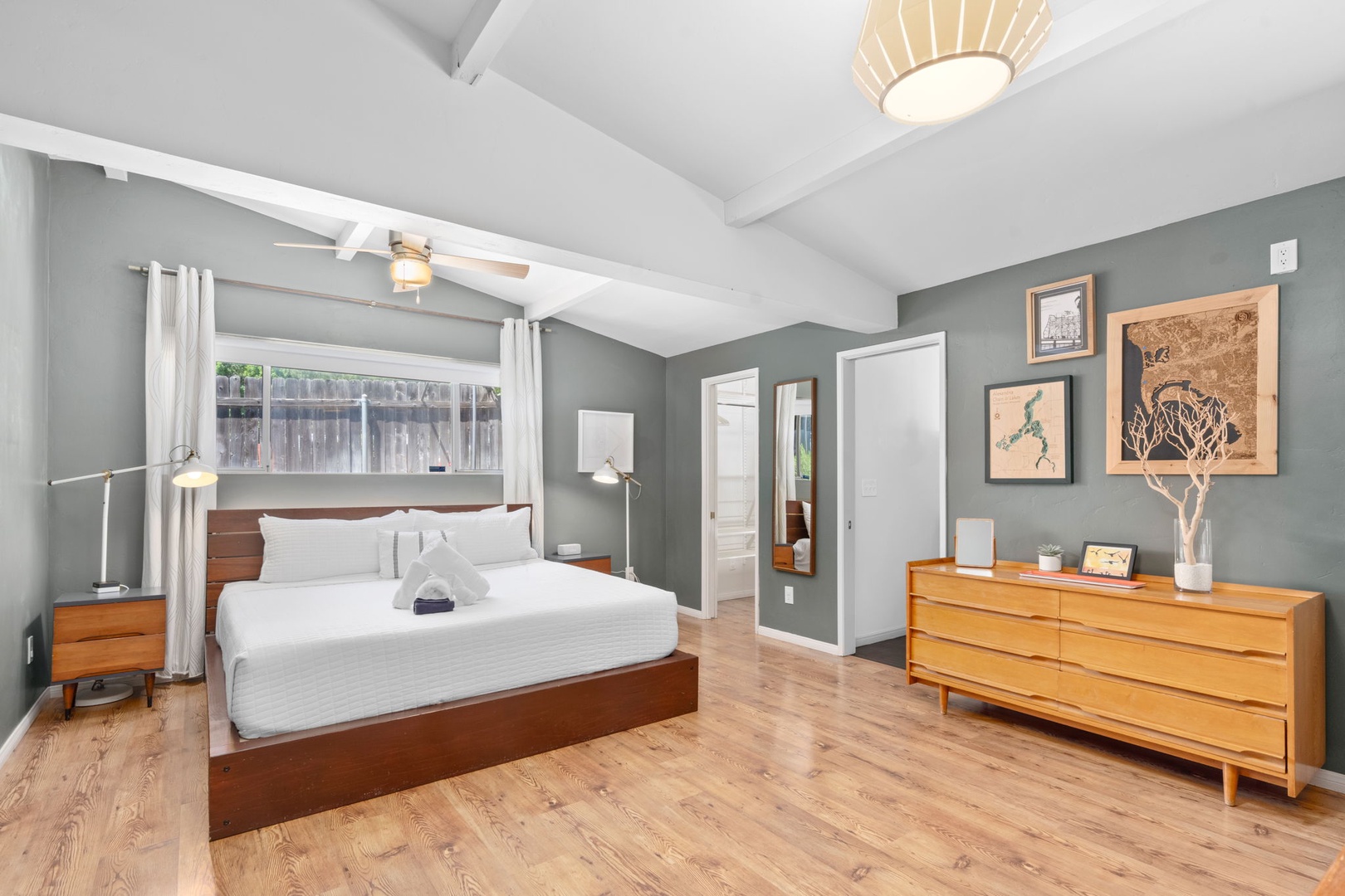 Sink into crisp white linens and wake to soft natural light in this serene primary bedroom. Mid-century wood furnishings and curated art warm calming sage walls, creating your peaceful retreat. Private ensuite bathroom and walk in closet.