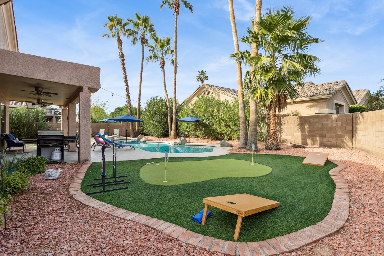 Desert oasis backyard features a sparkling pool, putting green, and shaded patio surrounded by towering palm trees.