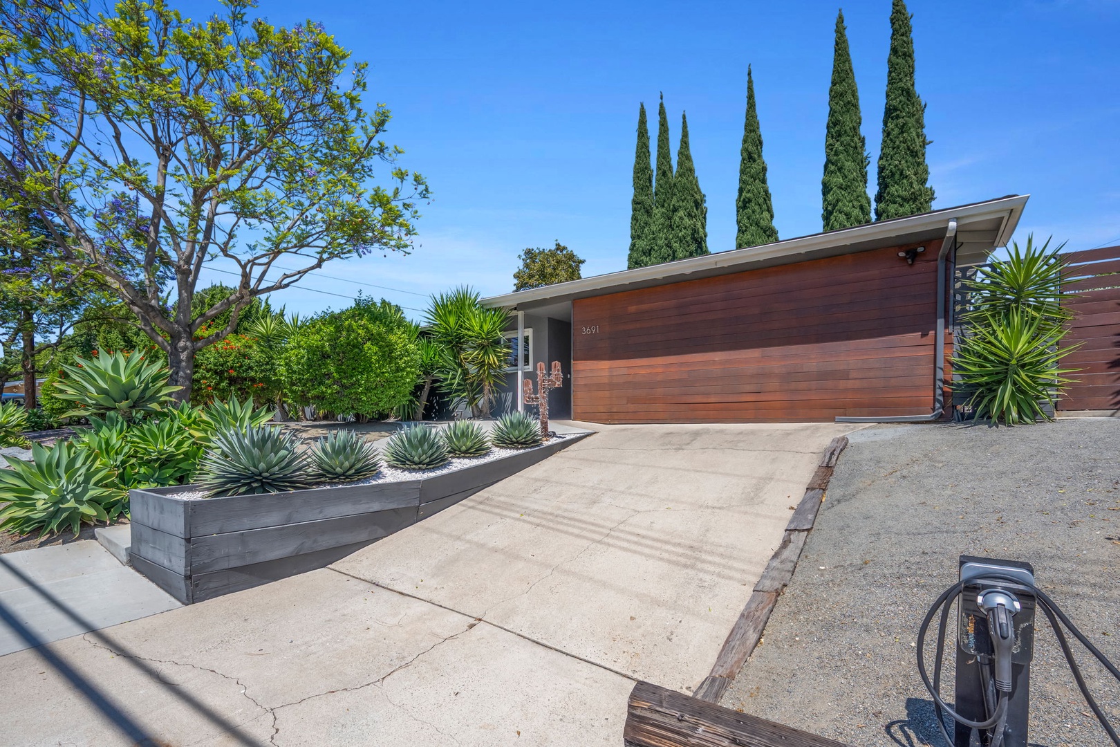 Mid-century modern home nestled among vibrant landscaping with towering cypress trees, native succulents, and colorful flowering bushes under sunny skies.