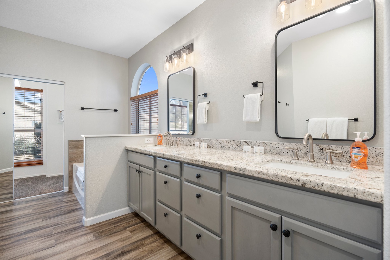 Primary suite bathroom features a spacious dual vanity with elegant granite countertops, modern fixtures, and plenty of natural light streaming through charming arched windows.