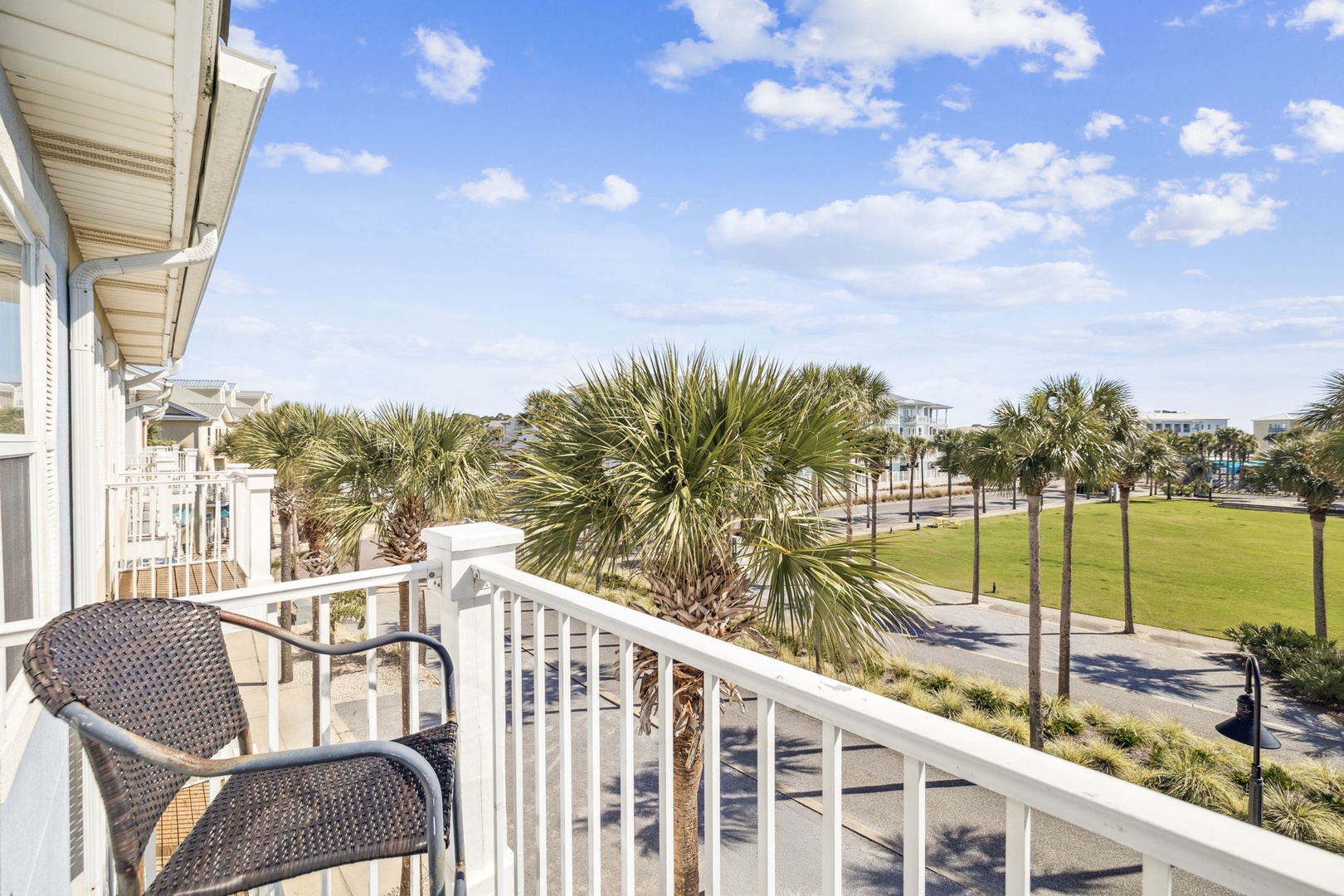 Coastal property balcony overlooking palm-lined green space and neighboring beachside community under sunny skies.