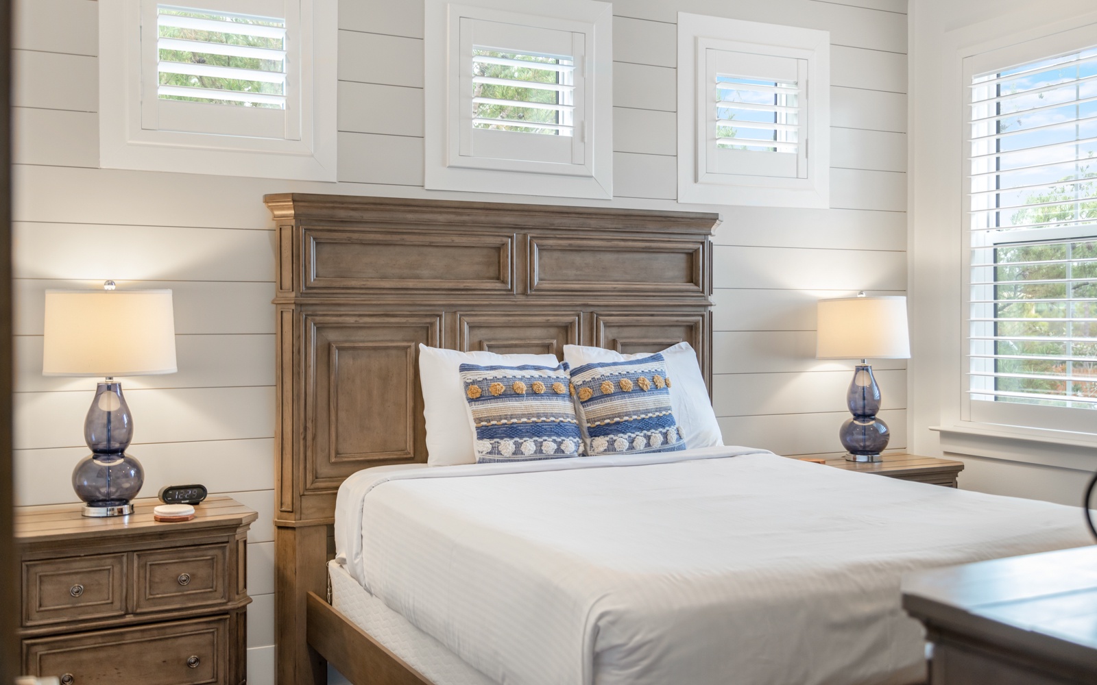Your peaceful bedroom sanctuary features crisp white linens and rustic wood furnishings, perfectly lit by natural light filtering through plantation shutters.