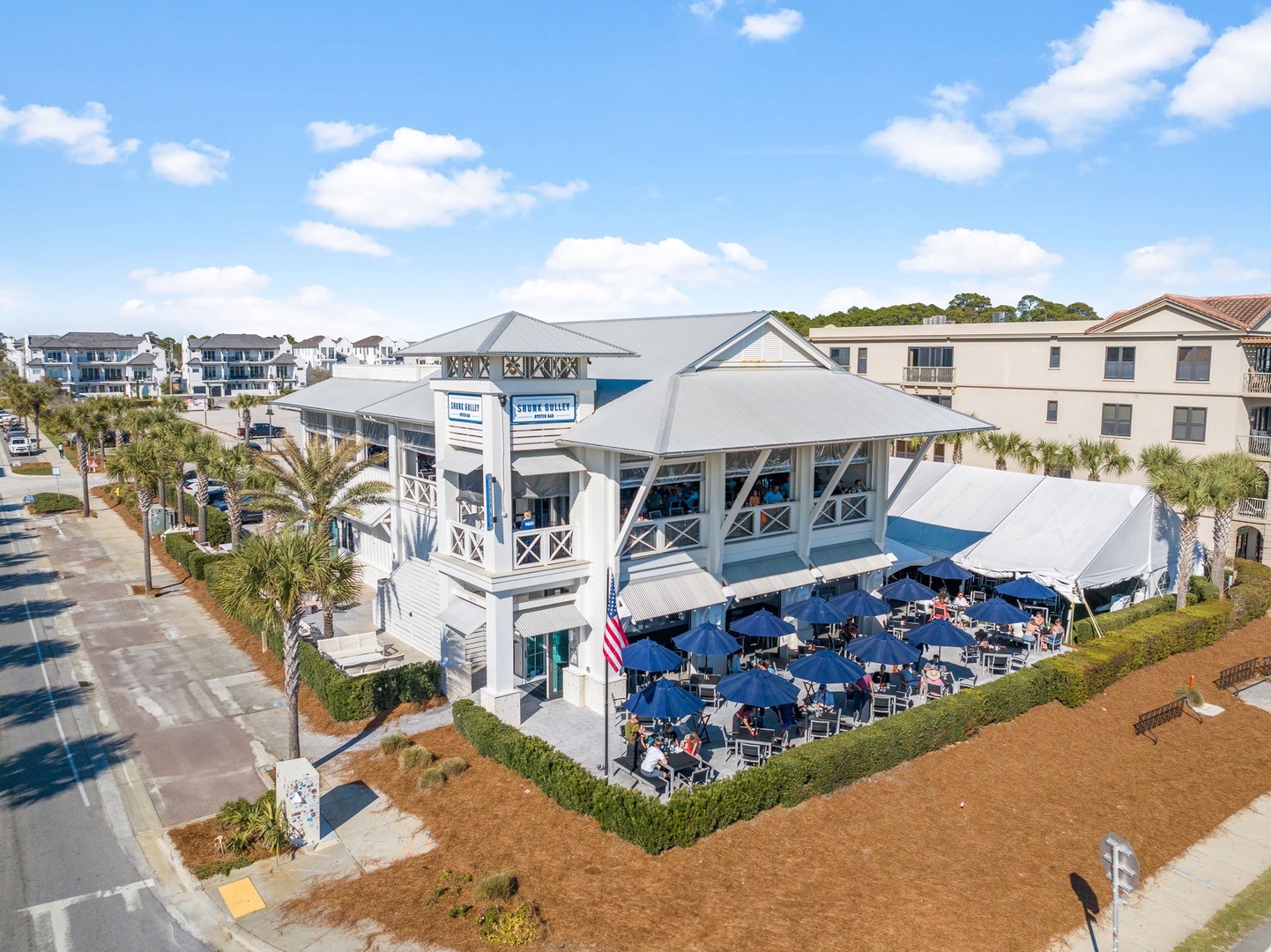 Aerial view of a charming coastal restaurant with outdoor seating area surrounded by palm trees and residential buildings.