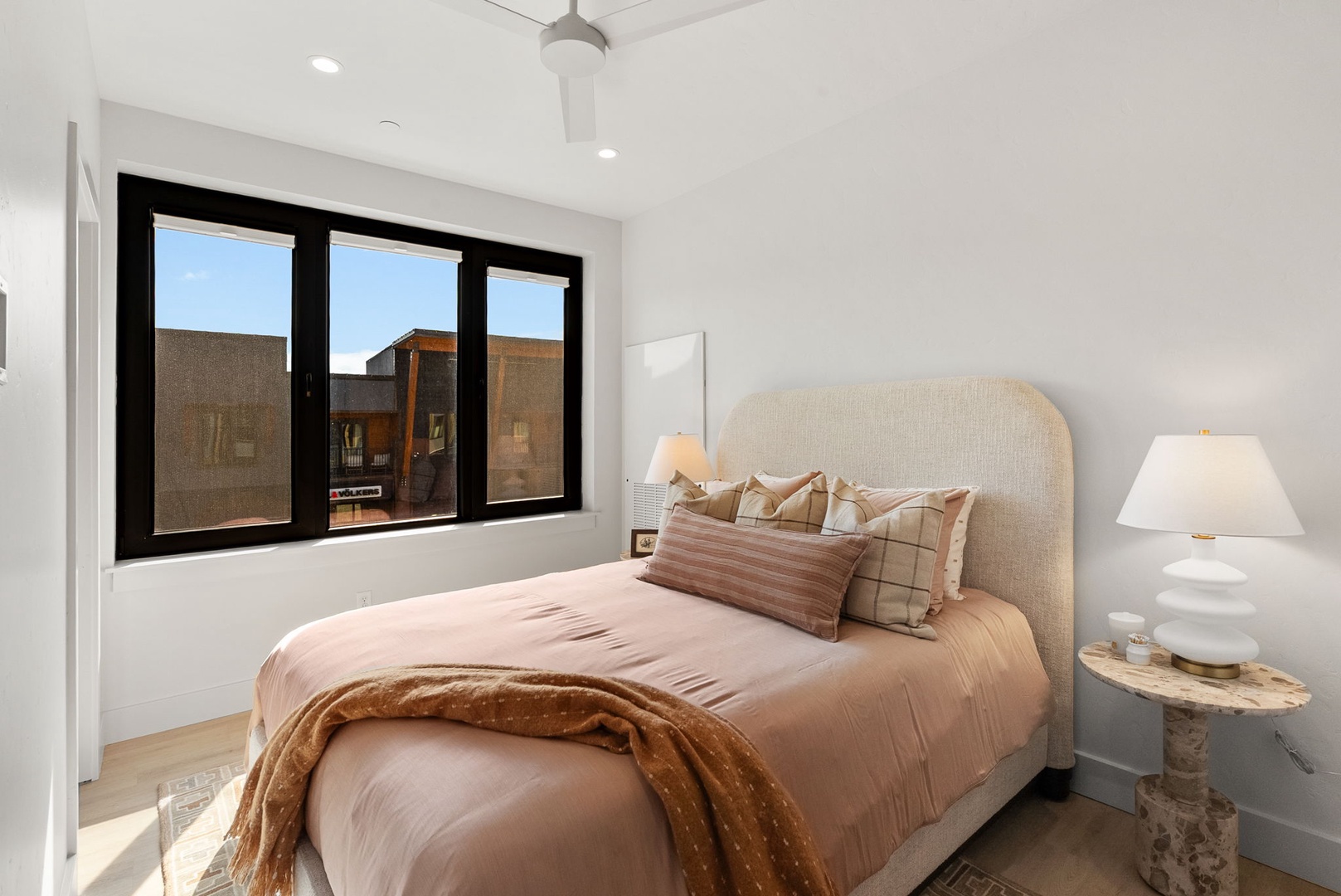 Your serene bedroom features a plush upholstered bed dressed in warm, inviting linens. Black-framed windows frame desert views while a ceiling fan keeps you cool after a day of exploring.