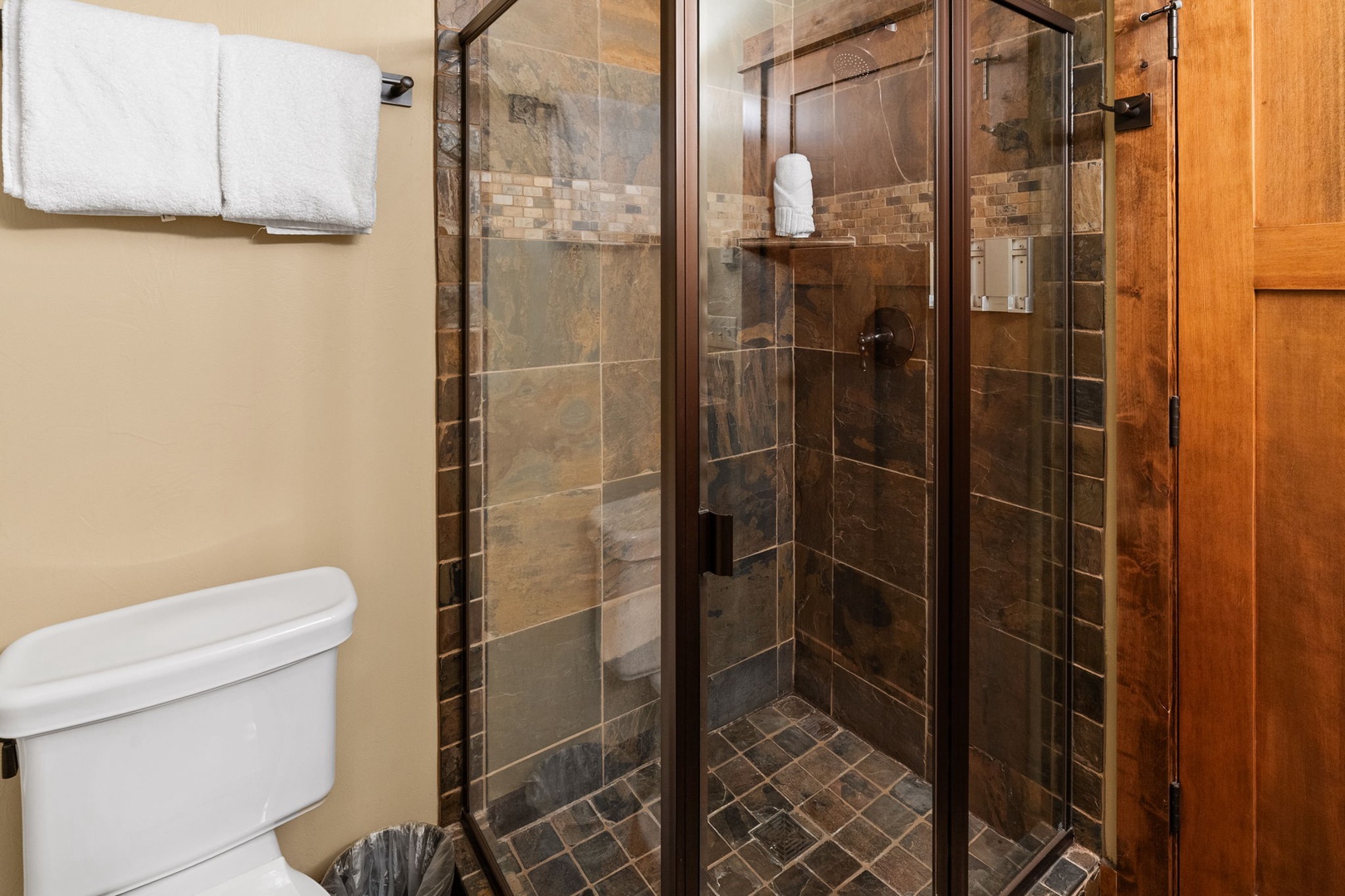 Step into your private bathroom with a beautifully tiled walk-in shower featuring natural stone tones and convenient built-in storage. Fresh towels await, and the rustic wood door adds mountain-lodge warmth.