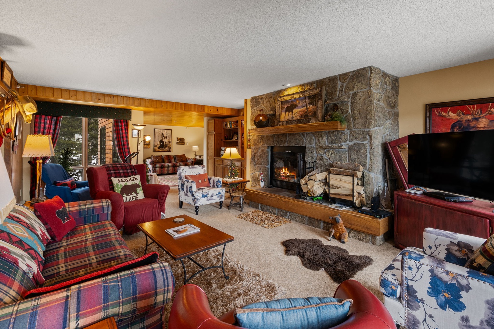 Cozy up by the crackling stone fireplace in this rustic living room, where plaid furnishings and warm wood create your perfect mountain retreat sanctuary.
