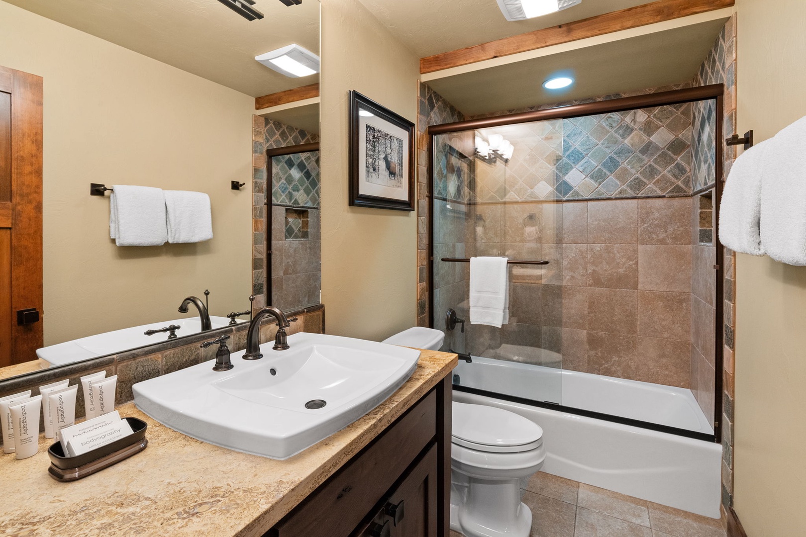 Step into your beautifully tiled bathroom with a modern soaking tub, warm wood accents, and plush towels awaiting your arrival. The glass-enclosed tub invites relaxation after a day of adventure.