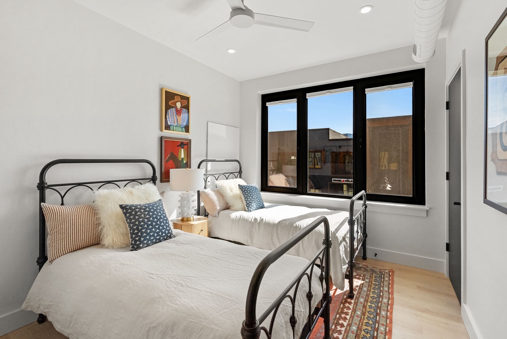 Relax in this bright, modern bedroom featuring two twin beds with vintage-style iron frames and a ceiling fan for your comfort. Large windows fill the space with natural light, creating a cheerful retreat.