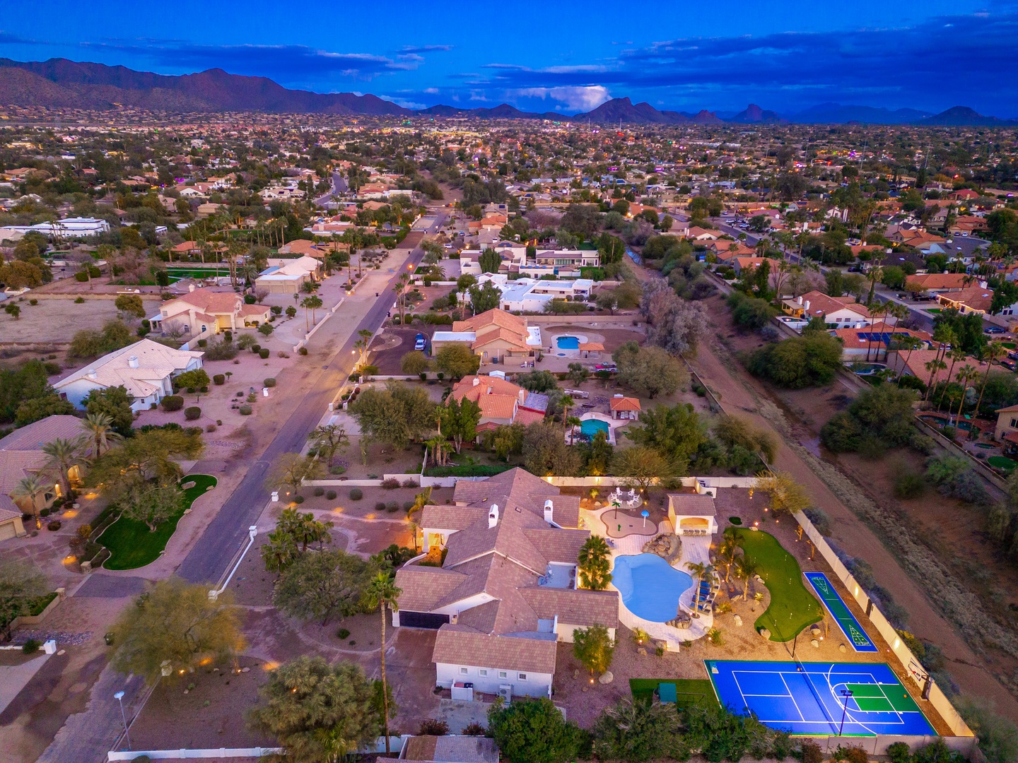 Aerial view of the property at dusk, nestled in a serene desert neighborhood with mountain vistas, private pool, and sport court—a scenic Arizona retreat.