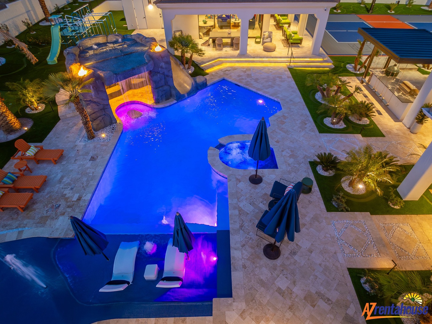 Stunning aerial view of luxury villa featuring illuminated pools, hot tub, waterslide, and pristine outdoor entertainment areas at dusk.