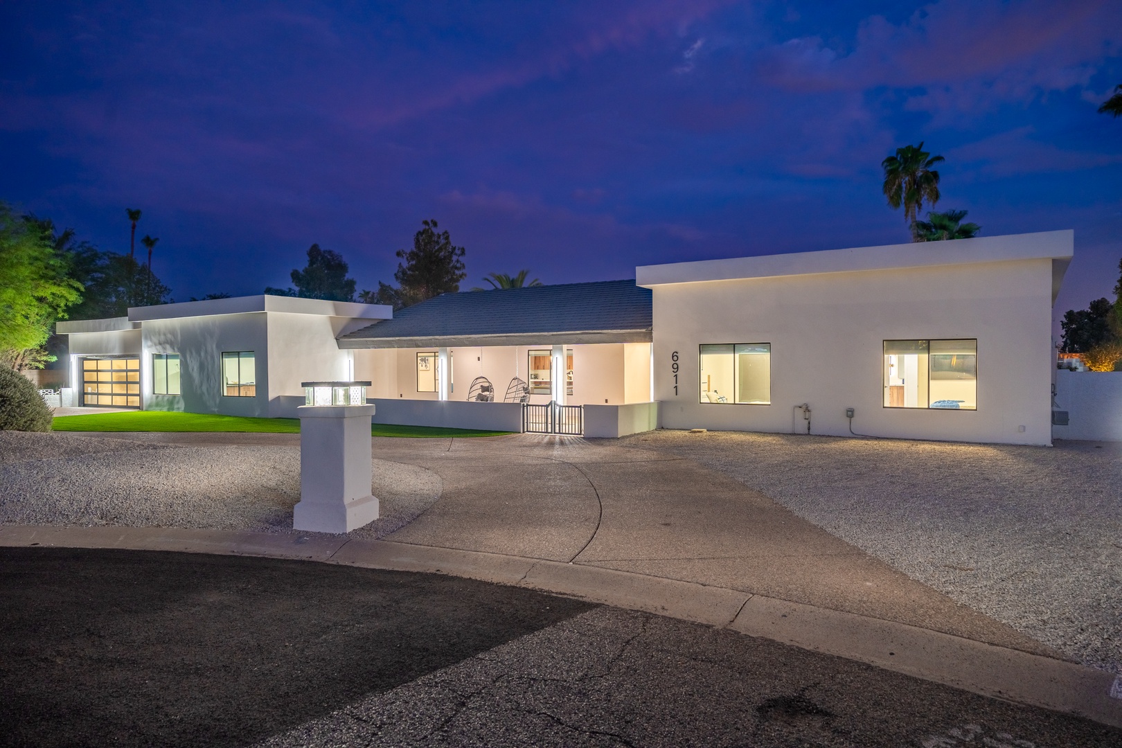 Modern single-level property featuring contemporary architecture, well-lit interiors, and manicured landscaping with palm trees set against a twilight sky.