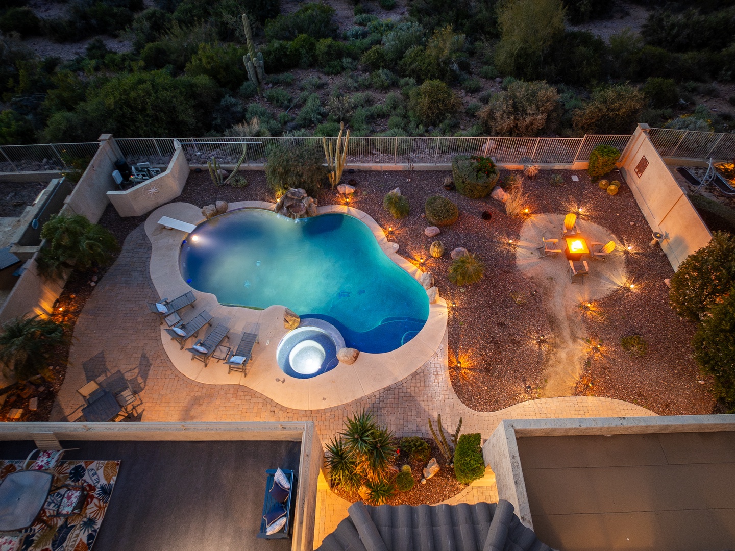 Aerial view of a serene desert retreat featuring a free-form pool, integrated hot tub, and illuminated patio area surrounded by natural Sonoran landscape and mountain views.