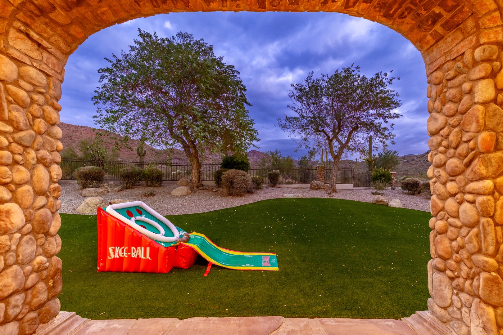 Step through the stone archway into your private backyard oasis—a lush lawn, colorful skee-ball slide, and desert mountain views await beneath twilight skies.