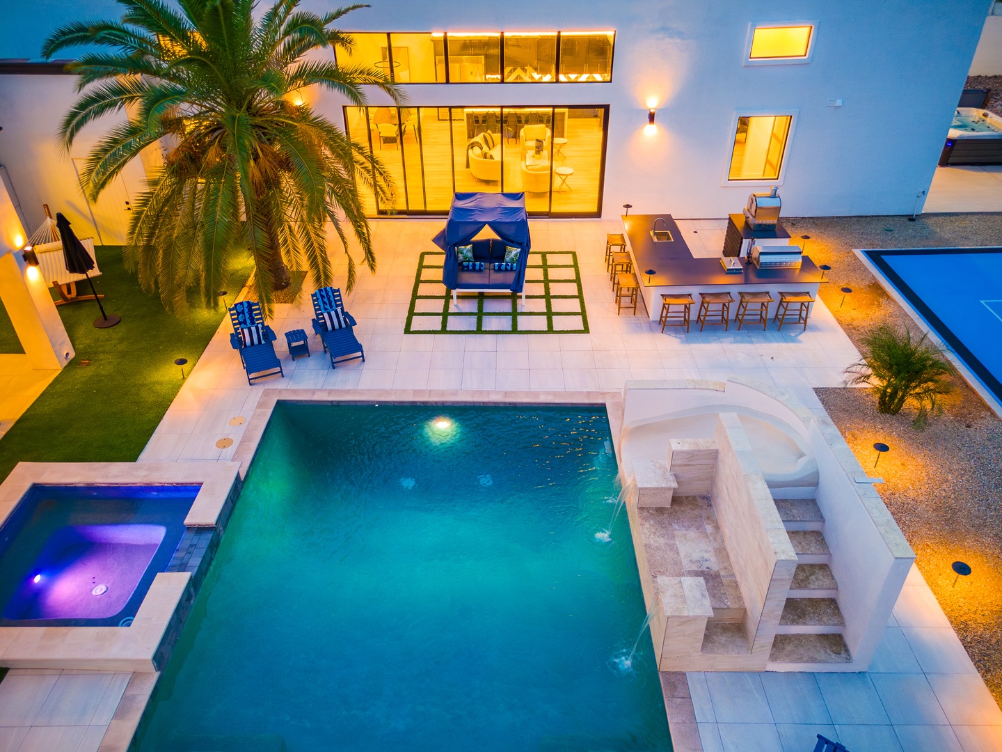Aerial view of a modern vacation villa showcasing a sparkling pool, hot tub, outdoor dining, cabana seating, and palm-lined patio at twilight—the perfect tropical escape.