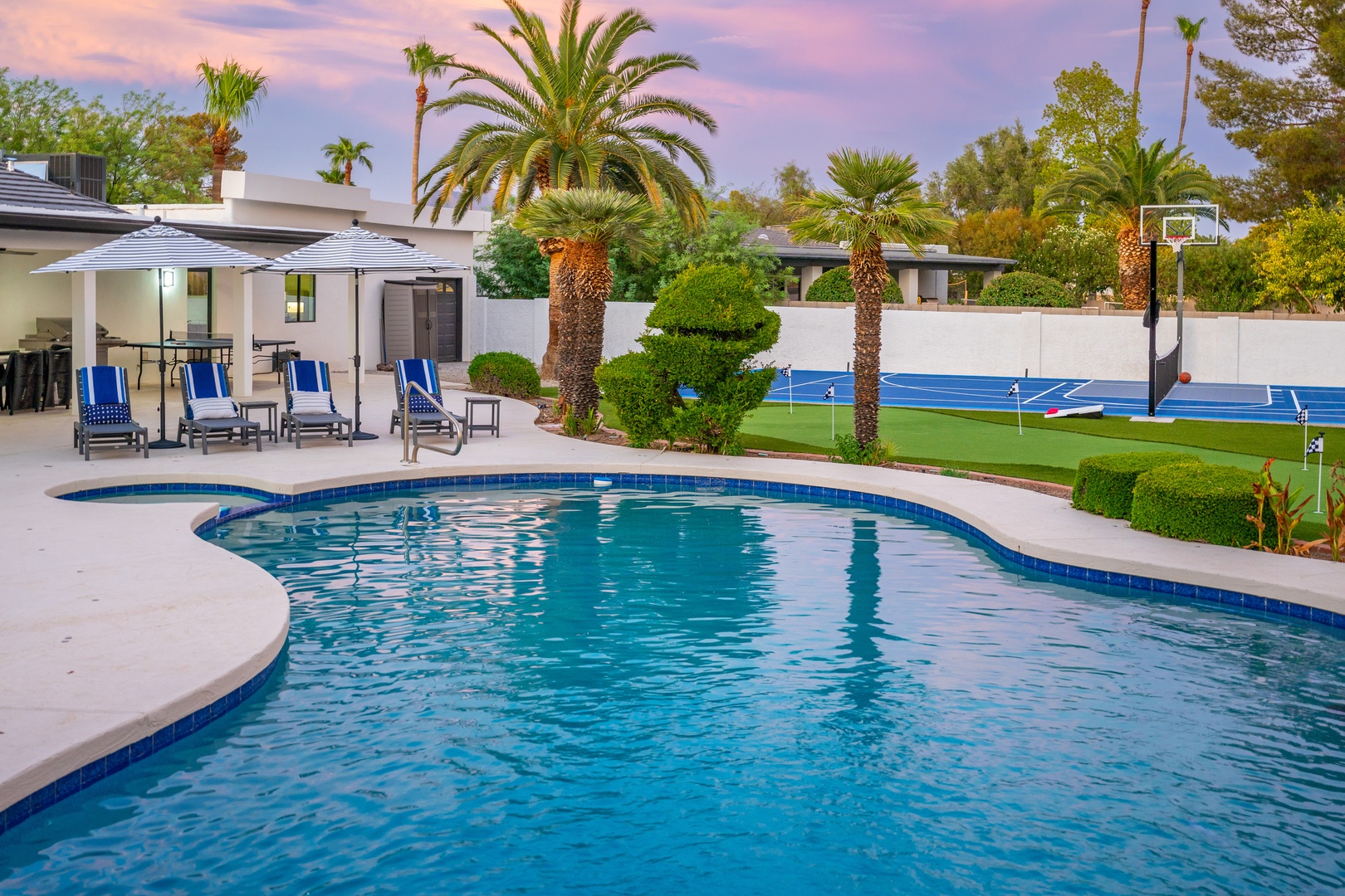 Your private oasis awaits with a sparkling pool framed by swaying palms, a shaded patio with lounge chairs, and a full-sized sport court for friendly competition under desert skies.