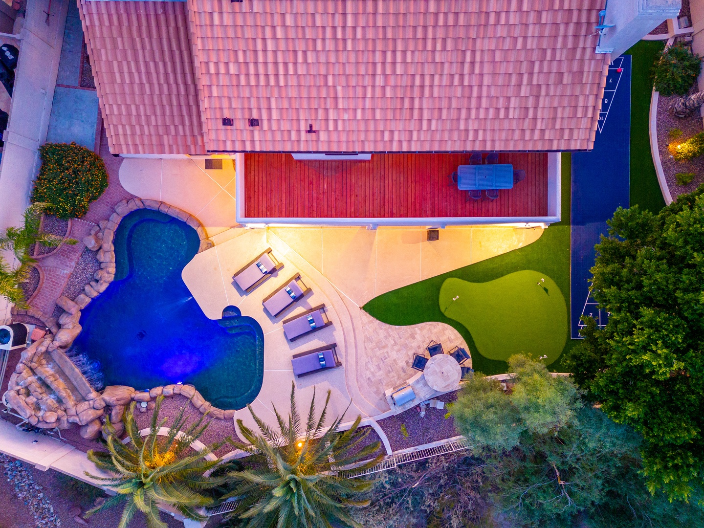 Aerial view showcasing the property's outdoor amenities including a curved swimming pool with rock features, poolside lounging area, putting green, and landscaped gardens with palm trees—all illuminated beautifully at dusk.