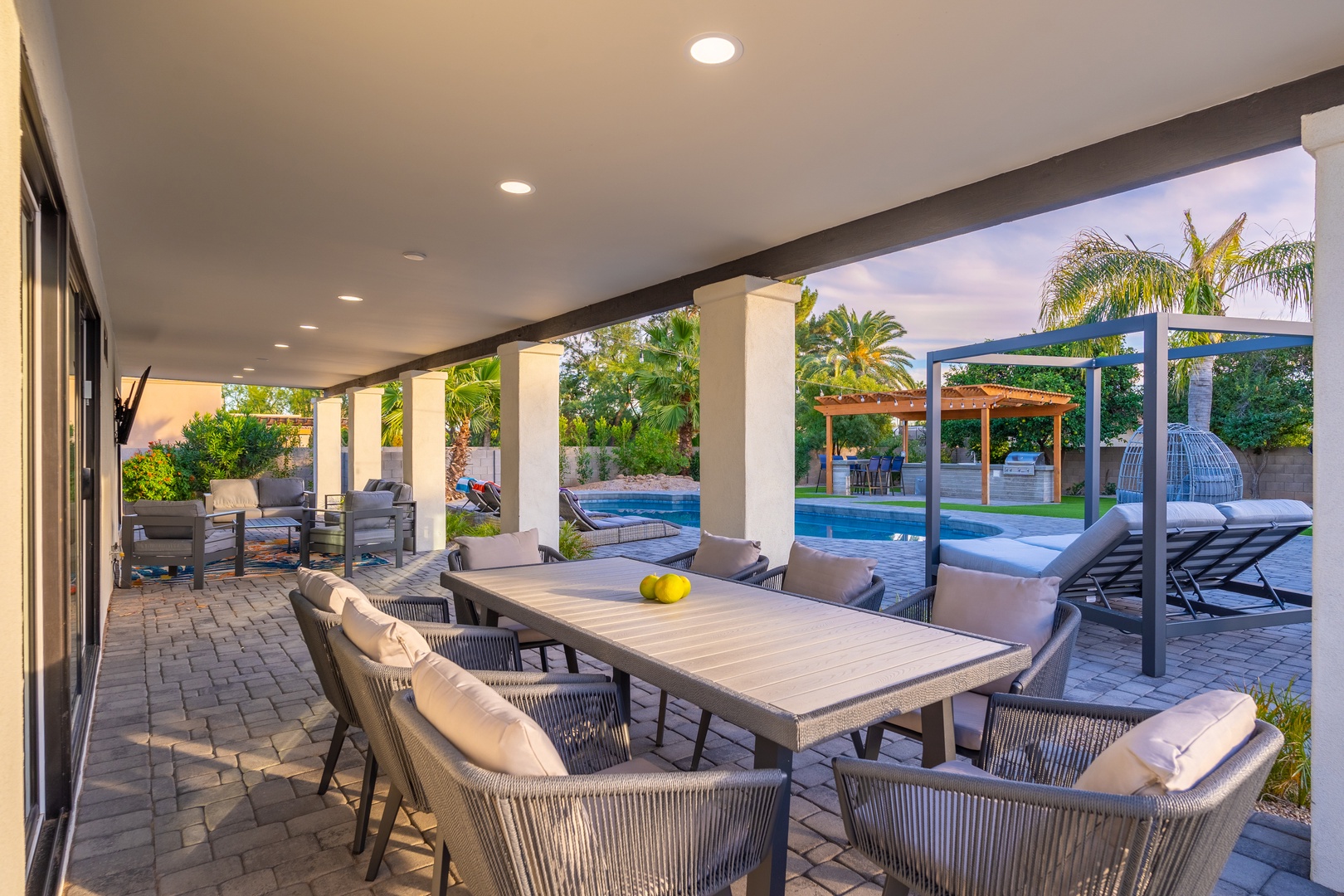 Covered patio with dining and lounge areas overlooks a sparkling pool, outdoor kitchen, and lush tropical landscaping, perfect for relaxed evenings or entertaining friends.